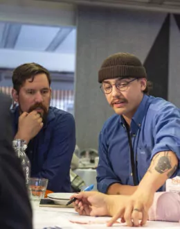 A group of people sitting at a table, collaborating. A man in a blue beanie and glasses points at a document while speaking, with others, including a man in a blue shirt and a woman with glasses, looking on.