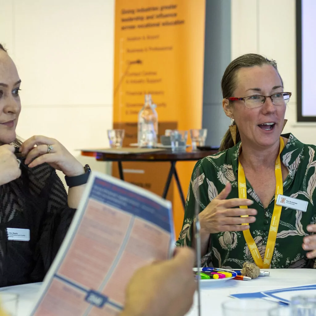 Two women sit together at a table discussing. One woman, wearing a black top and earrings, listens while the other, wearing a green floral shirt and yellow lanyard, gestures with her hands. A Ringa Hora banner is visible in the background.