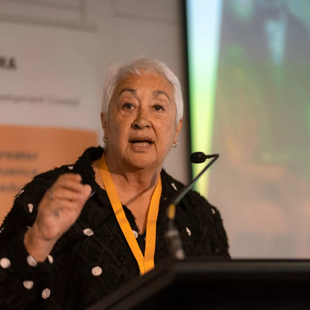 A woman with short white hair and a black polka-dotted sweater speaks passionately at a podium. She has a yellow lanyard and gestures with her right hand while addressing the audience.