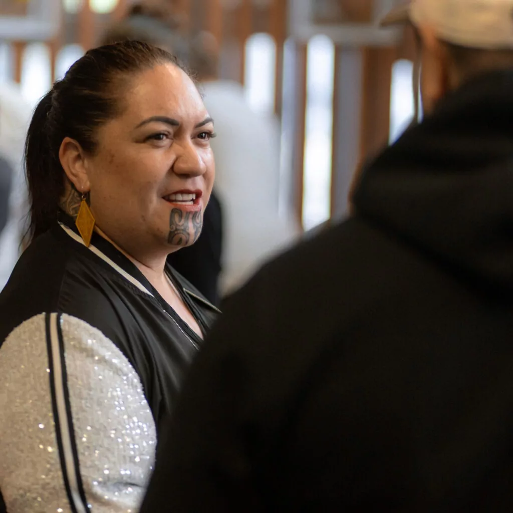 A woman with traditional moko kauae tattoo smiling and talking to someone off-camera.