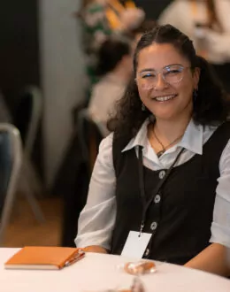 A seated woman with glasses smiling warmly at the camera during a gathering.