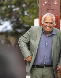 An elderly man in a light green suit and blue shirt smiles warmly while holding a walking stick. Behind him, a carved Māori post and greenery are visible, adding cultural significance to the setting.