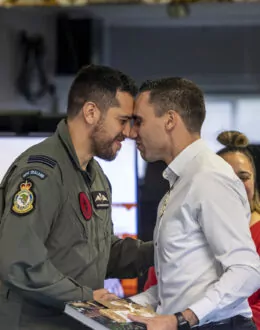 Two men share a hongi, a traditional Māori greeting, during a ceremony. One wears a military uniform with New Zealand Air Force insignia, and the other wears a white shirt. A woman in a red top is visible in the background, with a Ringa Hora banner to the side.