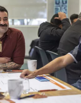Two men seated at a table during a discussion. One is smiling and wearing a red shirt with a pounamu necklace, while the other looks thoughtful in a navy polo. Papers and cups are on the table.