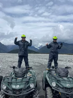 Two people standing on quad bikes. In the background are mountains.