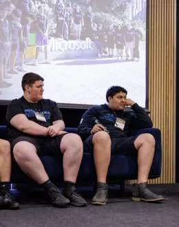 A group of people sitting down on a blue sofa. One person on the right is holding a microphone and talking to them.