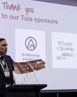 A side angle of a tane Māori (Māori man) doing a presentation. He is wearing a blue suit. In the background is a screen with different business logos.