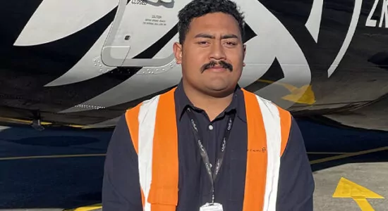 A person standing in front of a black and white plane. They are wearing a blue shirt and a bright orange Hi-Vas.