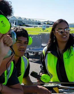 Three people on a runway strip. One person is pointing at something. They are wearing protective equipment. There are several planes in the background.