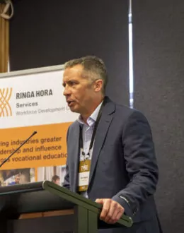 A side angle of a tane Māori (Māori man) giving a presentation from a podium. In the background is a RInga Hora banner.