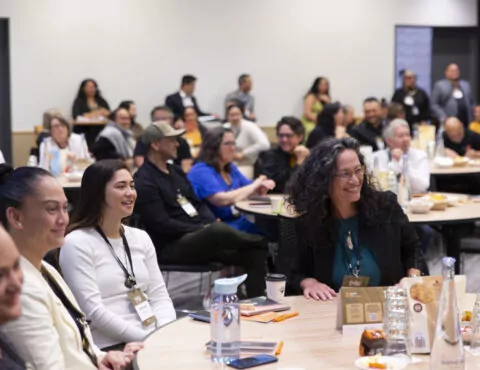 A conference room full of people smiling and sitting down at circular tables.