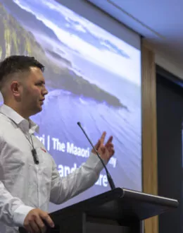 A side angle of a person presenting behind a podium. In the background is a screen wth an ocean and mountain range on it.