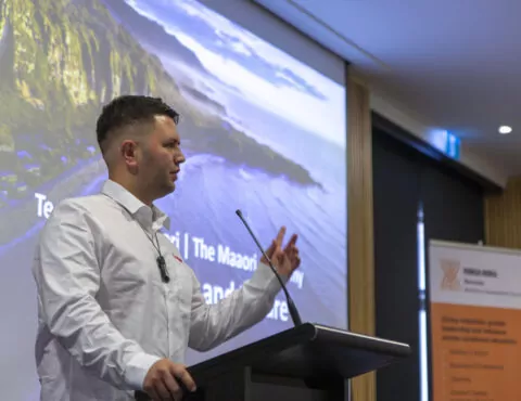 A side angle of a person presenting behind a podium. In the background is a screen wth an ocean and mountain range on it.