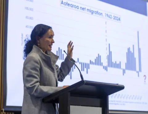 A side angle of a person standing behind a podium and doing a presentation. In the background is a screen that shows a bar graph of Aotearoa's migration rate.