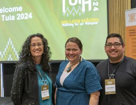 Three people posing together for the camera. In the background is a screen welcoming people to the 2024 Tuia event.