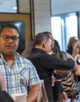 An over the shoulder close up of a person in a blue checkered shirt. In the background is a room full of people.