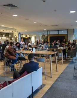 A conference room full of people sitting down. there is natural light shining through the big sliding doors to the entrance of the room.