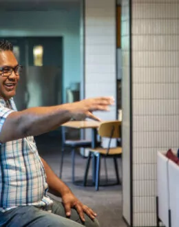 Side angle of person stretching his arm out and talking to someone. They are wearing a striped shirt. In the background are several other people sitting down.