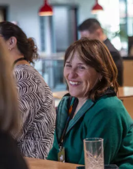 A close up of a person smiling and talking with someone at a table. They are wearing a green top.