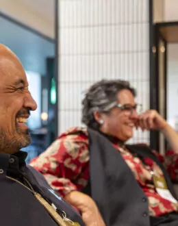 A close up side angle of a tane Māori (Māori man) laughing. There is a person in the background laughing as well.