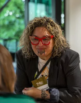 A close up of a wahine Māori (Māori women) standing at a table. She is wearing red glasses, and a black jacket.