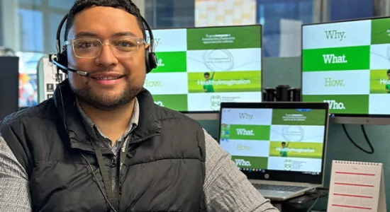 A person with short dark hair, glasses, and a headset, smiling while seated at a desk in a modern office environment. Multiple monitors in the background display green and white graphics.
