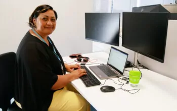 A woman sits at a modern office desk, typing on a laptop connected to multiple monitors. She wears a black blouse and yellow pants, with a blue ID lanyard around her neck. A green reusable coffee cup sits on the desk. She smiles warmly at the camera, with a professional and organized workspace behind her.